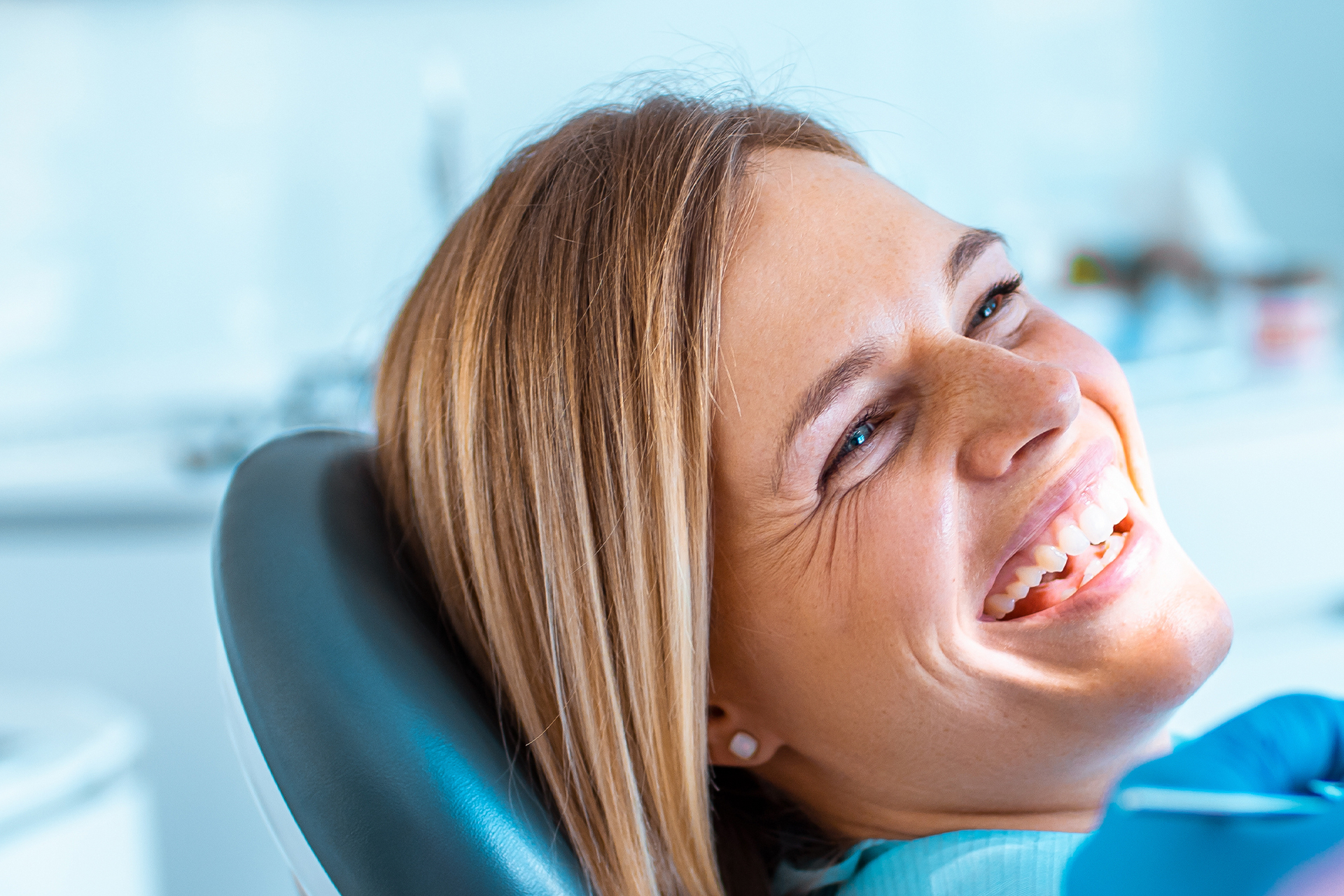 A woman sitting in a dental chair with a big smile on her face, wearing glasses and a blue shirt, appears to be having a dental appointment.