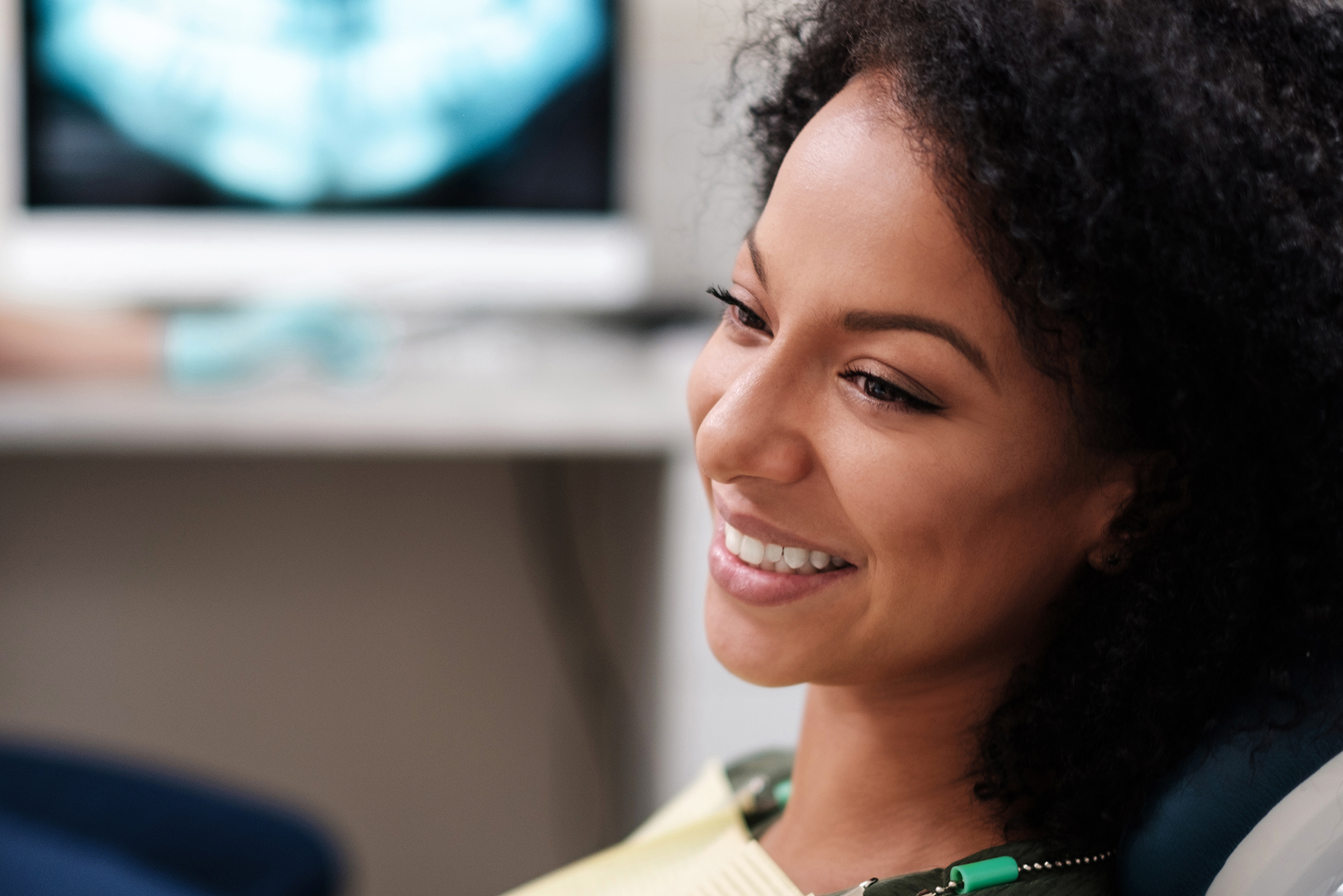The image shows a smiling woman with dark hair seated in front of a medical device, possibly a dental chair, with a bright background suggesting a professional healthcare setting.