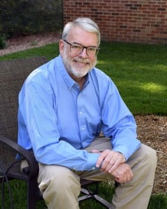 The image shows a man sitting on a chair outdoors, wearing glasses, with a relaxed posture, smiling slightly at the camera. He has gray hair, is dressed in a blue shirt, and appears to be posing for the photograph.