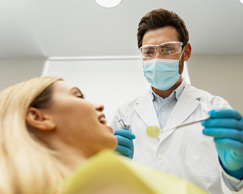 The image features a dental professional in a white lab coat performing an examination on a patient s teeth using a dental mirror, with the patient seated in the foreground and the dentist standing behind them.