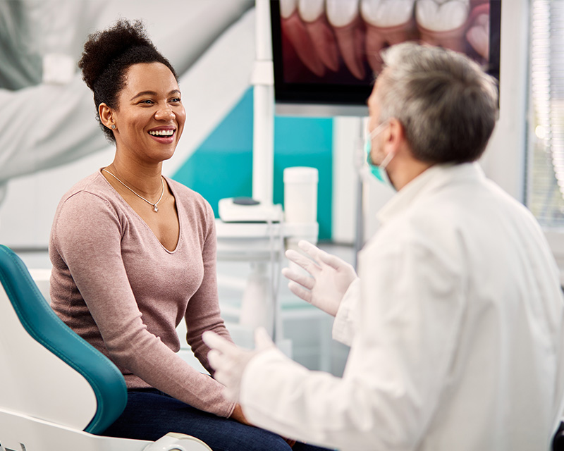 The image depicts a dental office setting with a woman sitting in a dental chair being examined by a dentist, who is standing and smiling at her.
