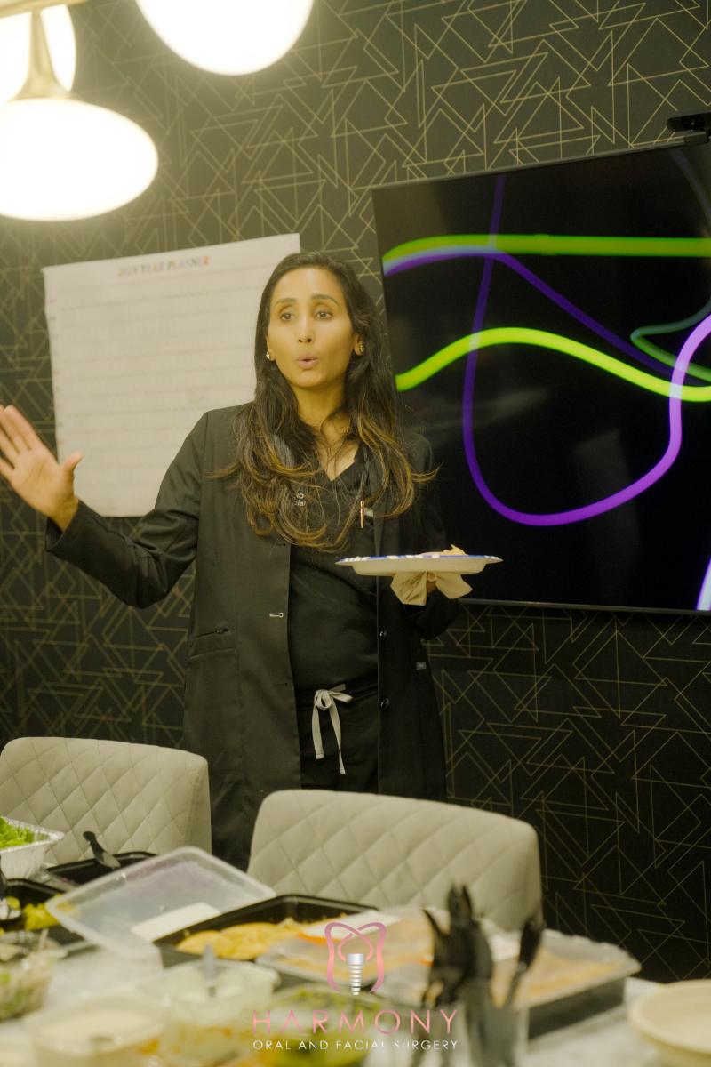 A woman stands at a conference table gesturing with her hands while addressing an audience.