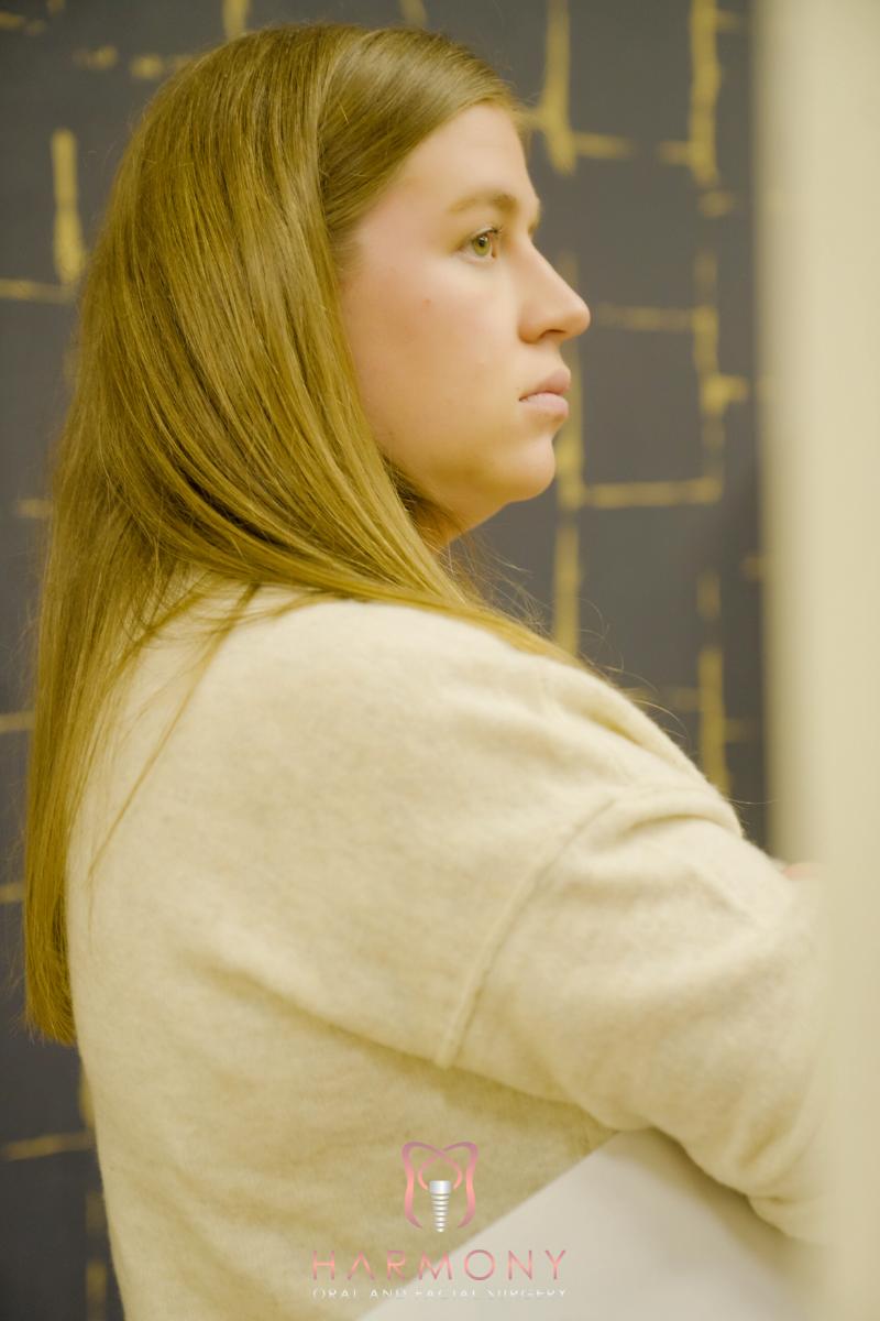 A woman with long hair stands in front of a patterned backdrop, facing slightly away from the camera.