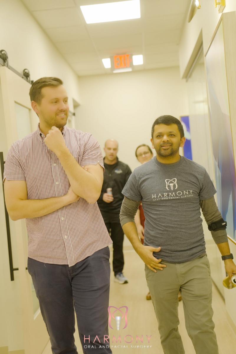 Two men walking side by side through an indoor space with a whiteboard and a door in the background.