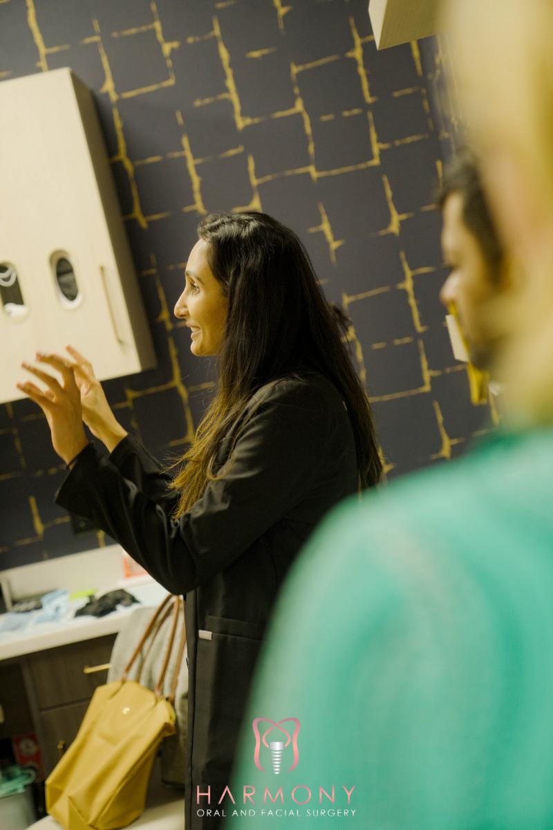 The image features two individuals standing in front of a backdrop with patterns, where one person appears to be speaking or gesturing towards something out of frame.