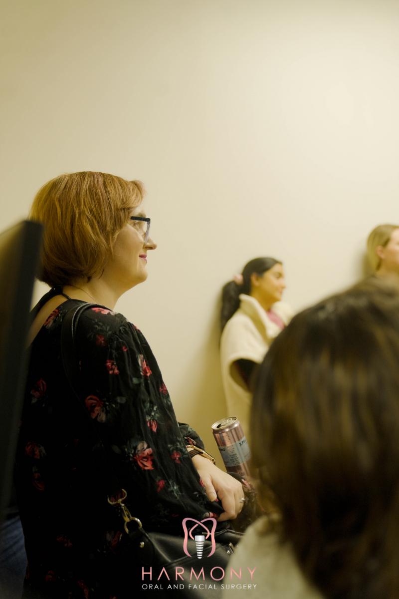 The image shows a person sitting down with a group of others, looking towards the camera with a slight smile. They are wearing glasses and have short hair. The setting appears to be an indoor space, possibly a room for presentations or meetings, given the presence of other individuals who seem to be engaged in conversation.