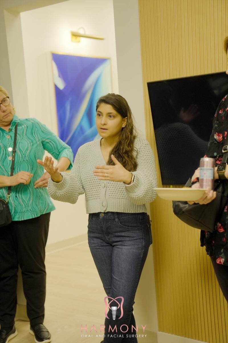 A woman standing in a room with two other individuals, gesturing with her hands while speaking, against a backdrop of a wall-mounted screen displaying an abstract design.