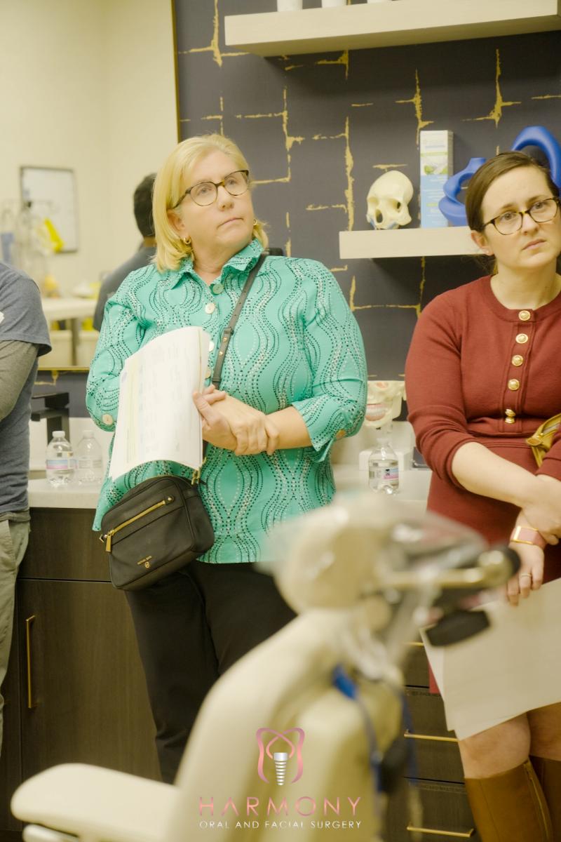 The image shows two women standing in front of a dental office counter with various items on display, including bottles and a model mouth. One woman appears to be holding a clipboard and is wearing glasses, while the other has a purse over her shoulder and is looking at something off-camera. They are both dressed in casual attire and seem to be engaged in conversation or activity related to their visit to the dental office.