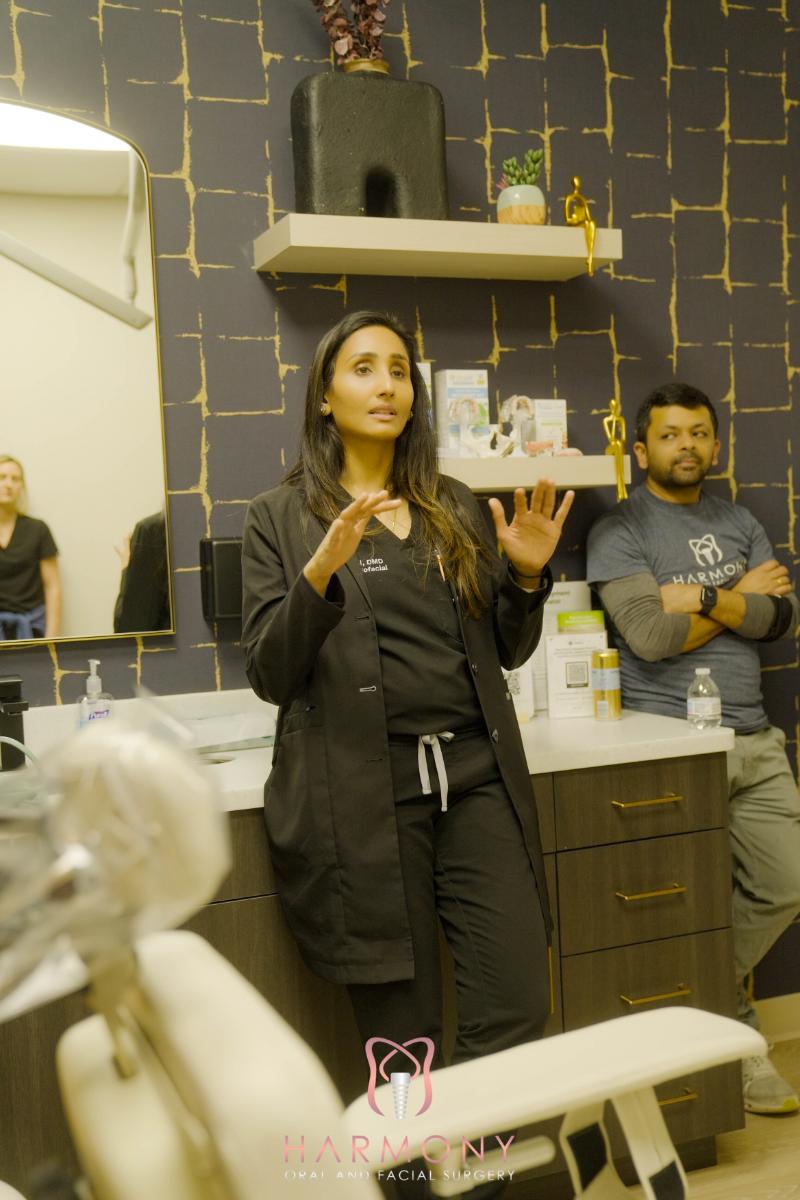 A woman stands in front of dental equipment, gesturing with her hands, while an observer watches from behind; both are in a dental office setting.