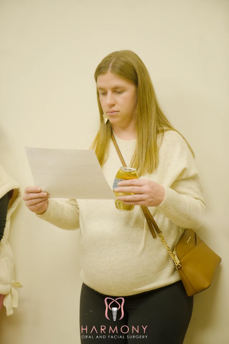 A woman standing against a white wall holding a piece of paper, reading from it.