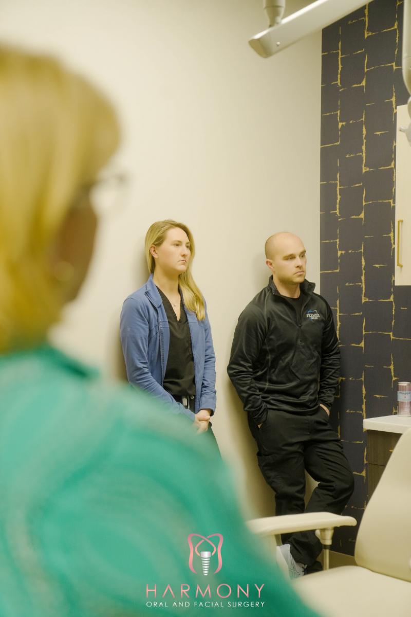 This image depicts two individuals standing in front of a blue wall with a black border, observing something on a screen, with a third person seated behind them. They are indoors, possibly in an office setting, and there's a window with blinds partially drawn to the left.