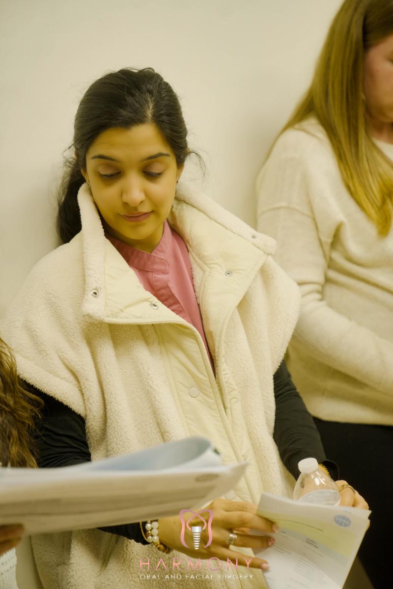 In the image, there is a woman seated on a chair in an indoor setting, looking down with a focused expression, surrounded by other people who appear to be engaged in different activities.
