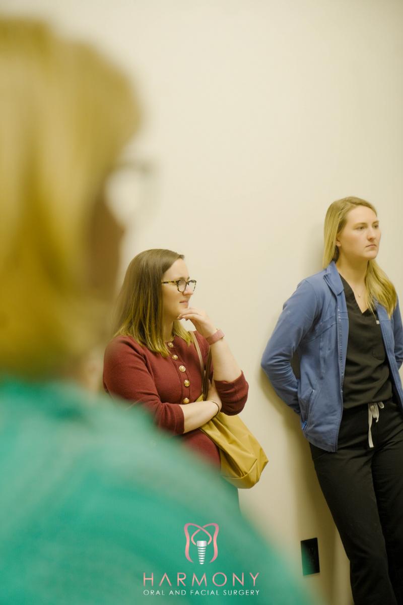 The image shows two individuals looking at something off-camera while standing near a wall with a sign. One person is wearing glasses, and they are both dressed in casual attire.
