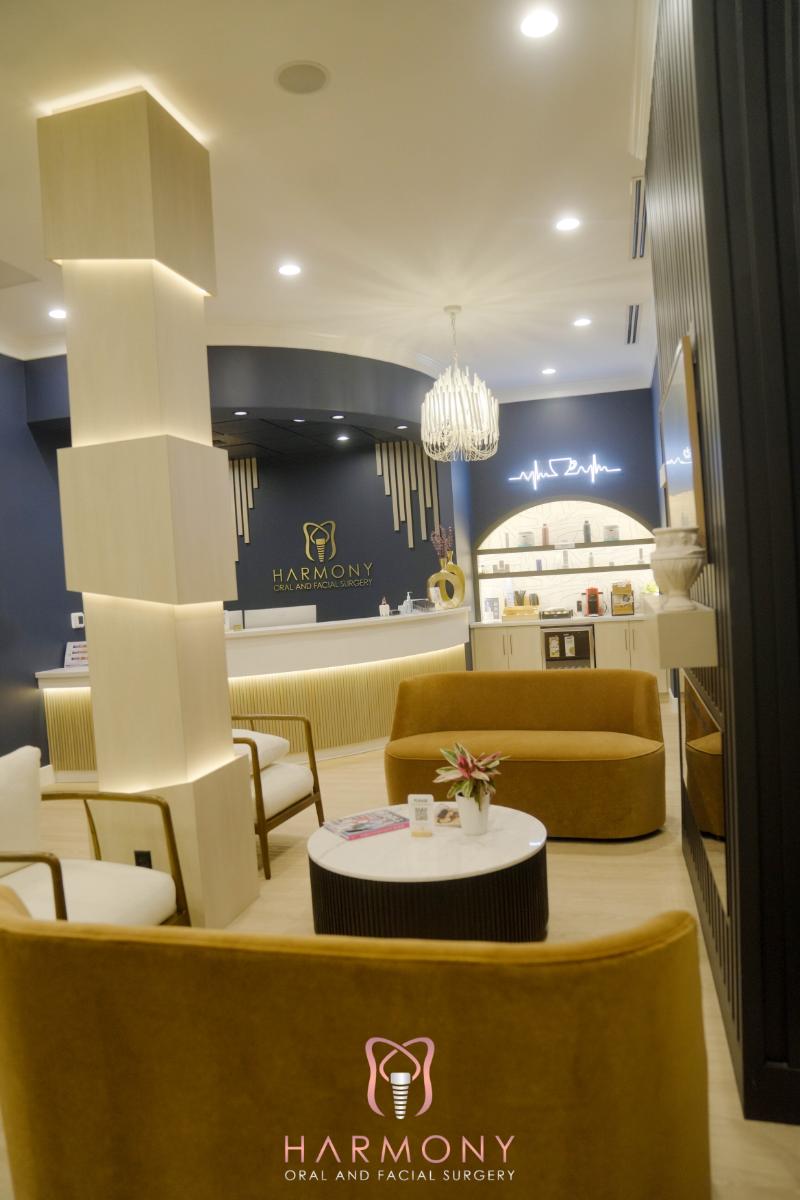 The image shows an interior space with a modern design, featuring a comfortable seating area with beige upholstered furniture, a bar counter with stools, and decorative elements such as plants and wall art.