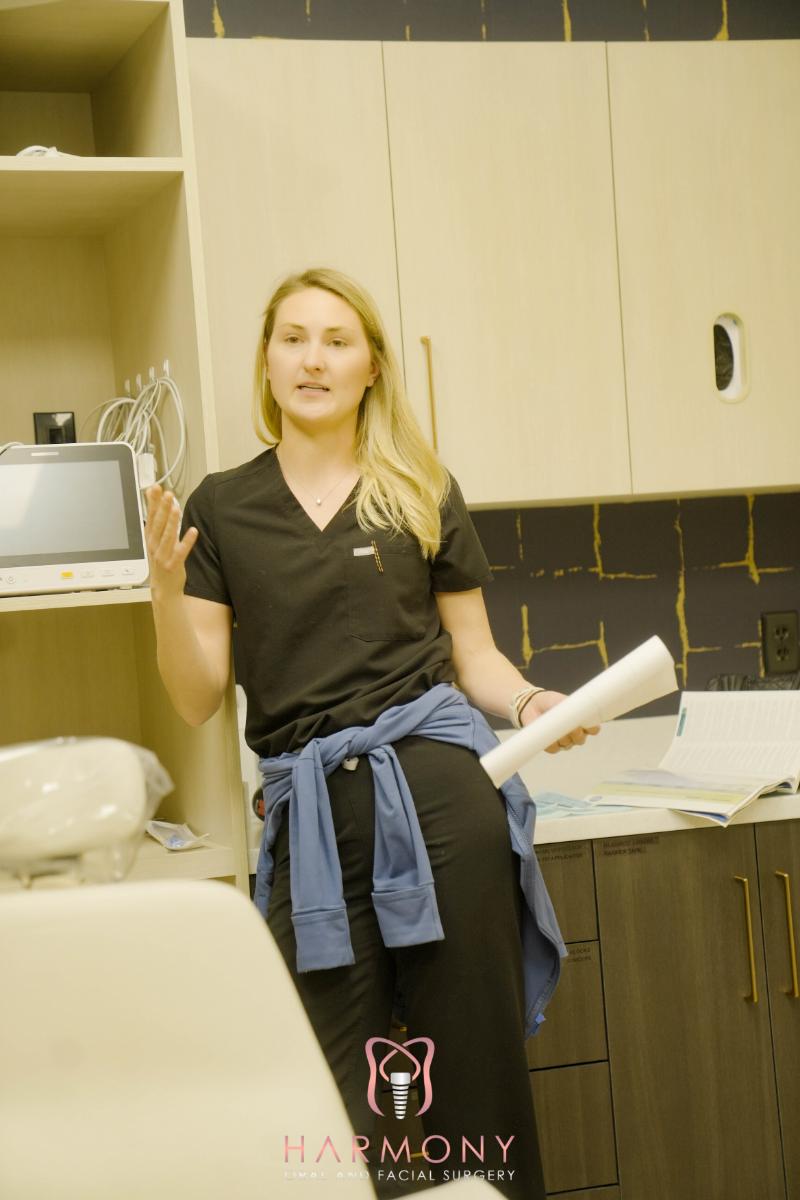 A woman in a blue shirt and black pants sitting in an office chair with her hand on her chin, gesturing while talking.