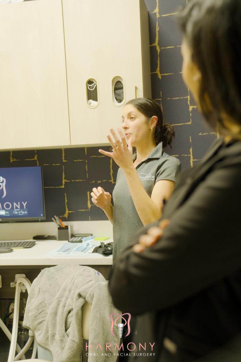 A woman standing at a desk with a laptop, gesturing and speaking to two individuals who are attentively watching her.