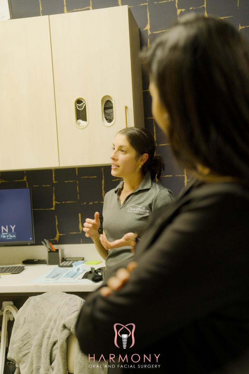 A woman stands at a desk with her hands gesturing while another person watches her from behind.