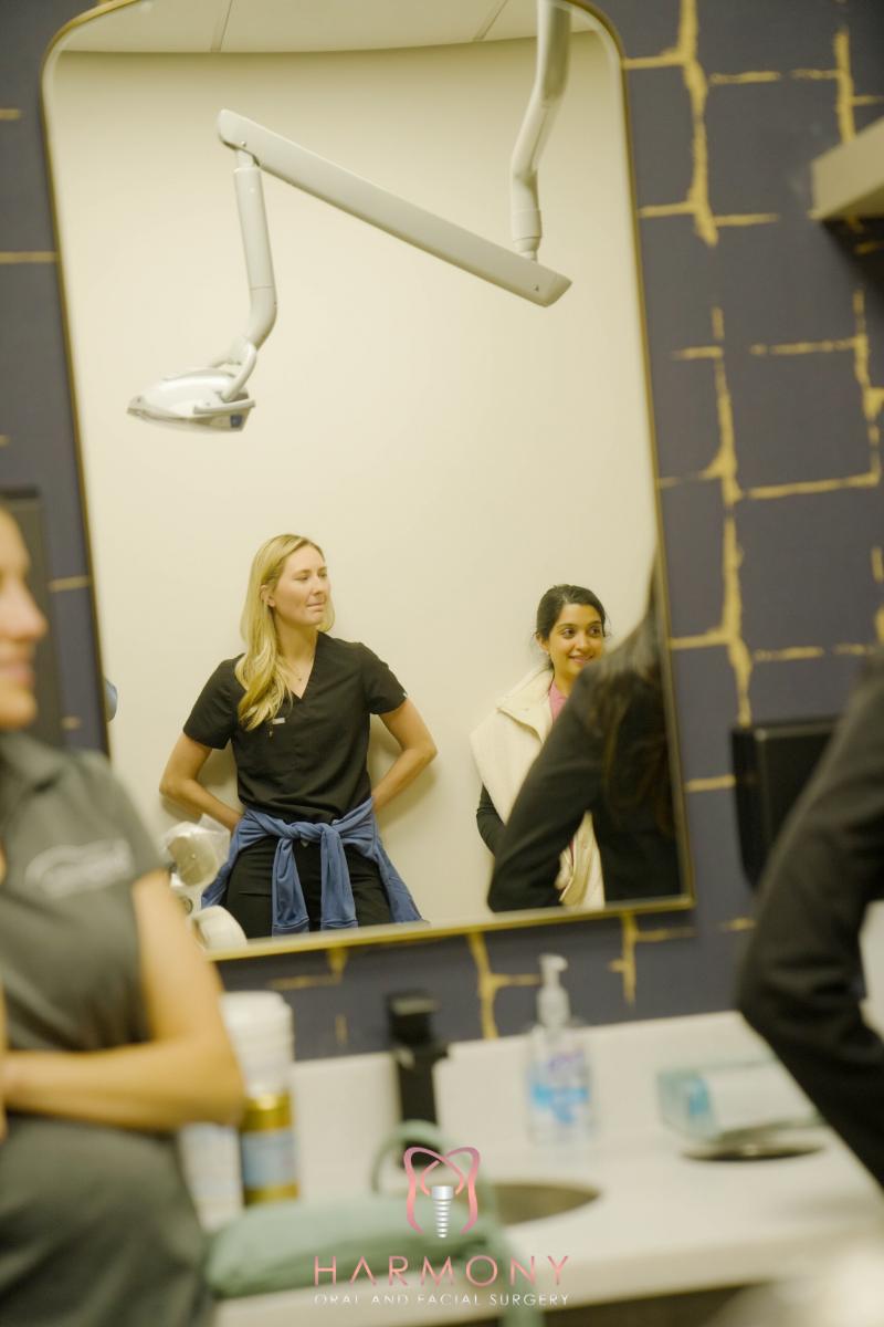 The image shows a woman standing in front of a mirror with another person, both in a room that appears to be a salon or barber shop, with various grooming tools visible in the background.