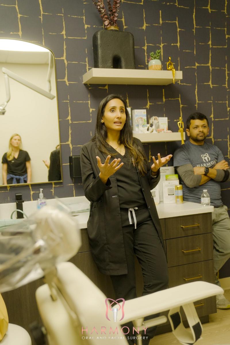 The image shows a woman standing in front of a dental chair in a dental office, gesturing with her hands while addressing an audience, with another person observing from behind.