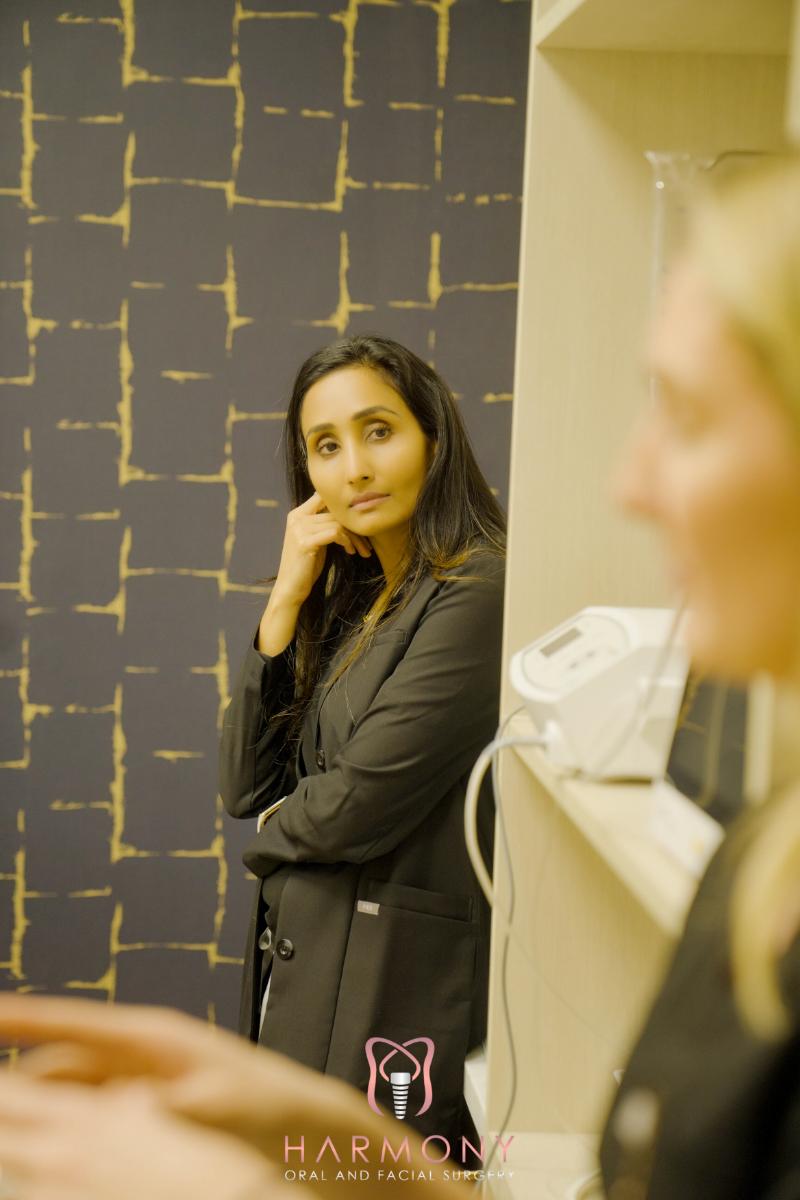 A woman stands in front of a counter, looking to her right with hands clasped together, while another person appears to be seated behind her, facing away from the camera.