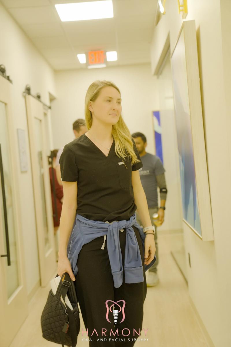 The image shows a woman walking down a hallway inside a building, wearing a black top and carrying a handbag, with a casual expression on her face.