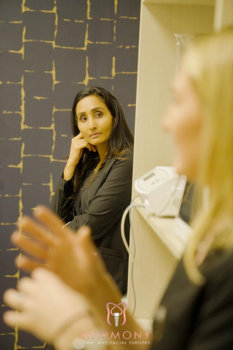A woman stands in front of another woman who is looking at her with a thoughtful expression. Both women are indoors, and one is holding a clipboard.