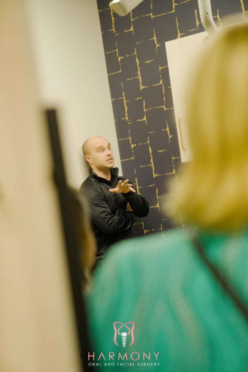 The image shows a man standing in front of an art piece, looking towards a woman who is also present in the scene. He appears to be gesturing with his hands.