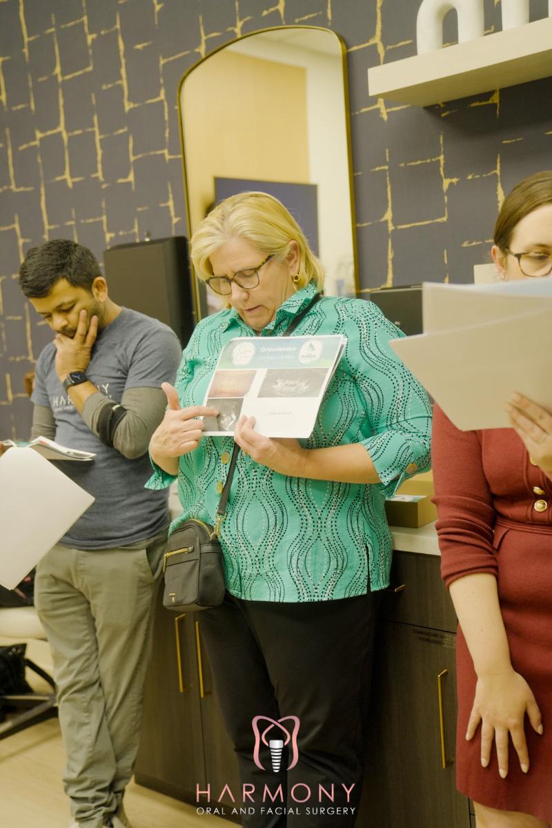 The image shows a woman standing at a counter with papers in front of her, holding a booklet, while two other people are present in the background. They appear to be in an interior setting, possibly a store or office, with a desk and various items on it.