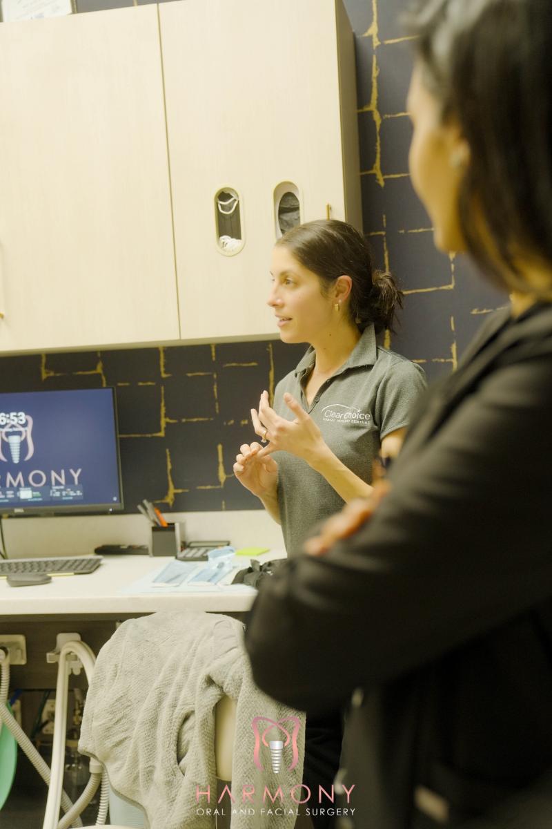 A woman standing next to a desk with a computer monitor, gesturing with her hands while speaking, in front of two women observing her.