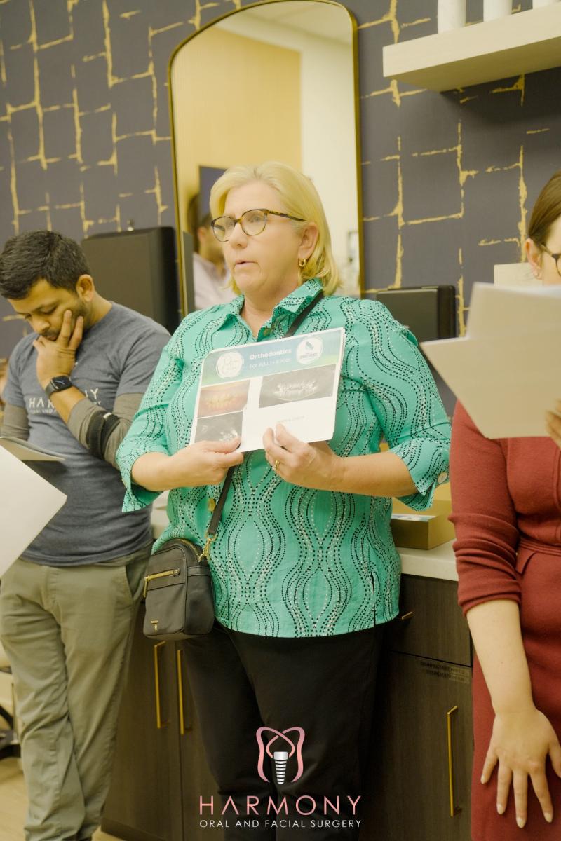 The image shows a woman standing at a counter with a sign, appearing to be presenting or explaining something, possibly in an educational or retail setting.