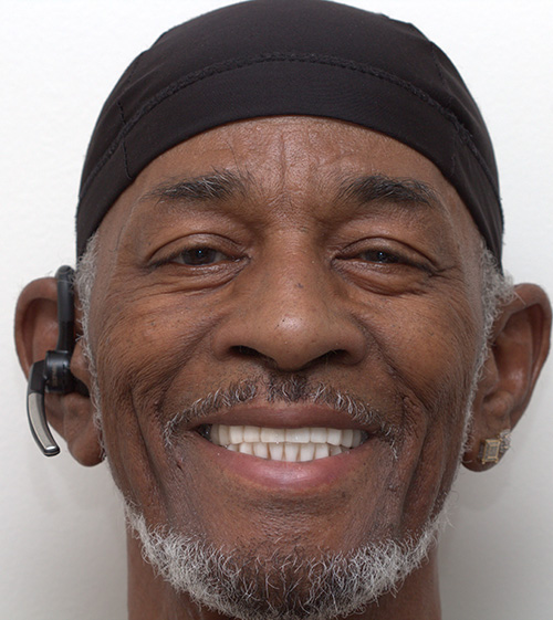 The image shows a man wearing headphones around his neck, smiling broadly at the camera with his eyes closed, against a plain background.